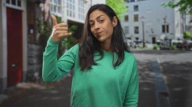 Genç bir kadın, yeşil kazak ve hafif göz kırpma giyerek, şehir caddesinde yükseltilmiş el hareketleriyle bana el hareketi der..