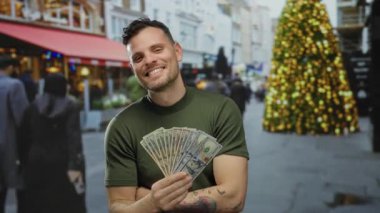 Bize dolar tutan genç adam, arka planda neşeli bir noel ağacı olan şehir sokaklarında gülümsüyor, canlı bir şehir atmosferinde Amerikan paralarını sergiliyor..