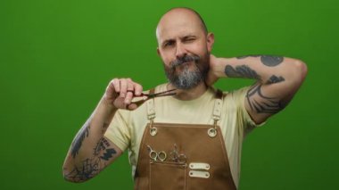Dövmeli sakallı adam elinde makasla yeşil arka plana önlük takıyor. Yorgunluğunu ve düşüncelerini dile getiriyor..