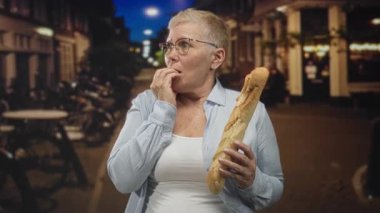 Elden çeneye ve çatık kaşlı, gözlüklü ve açık tişörtlü bir gece sokakta elinde baget tutan kadın; kentsel korku açlığı.