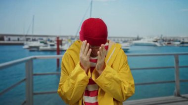 Sarı ceketli, kırmızı bereli genç adam açık hava altında teknelerin yanında el ele tutuşuyor..