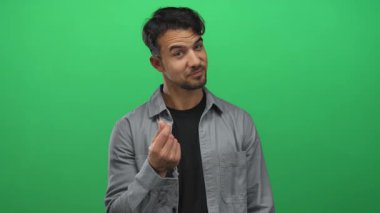 Genç İspanyol asıllı bir adam, yeşil bir arka plana parmaklarıyla para hareketi yapıyor. Günlük kıyafetleriyle neşeli ve kendinden emin bir tavır sergiliyor..