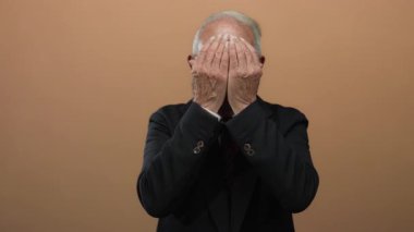 Takım elbiseli kıdemli adam, yalnız kahverengi arka planda şaşkınlığını dile getiriyor. Duygularını ve iş kıyafetlerini vurgulayarak yüzünü ortaya çıkarıyor..