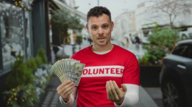 Latin kökenli genç bir adam Amerikan banknotlarını elinde tutuyor. Kırmızı gönüllü gömleği giyip el hareketi yapıyor..