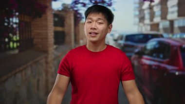 Kırmızı gömlekli genç adam şehir sokaklarında büyük bir jest yapıyor. Arka planda arabaları var. Heyecan ve nişan gösterisi yapıyorlar..