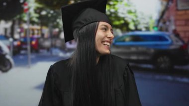 Bir şehir caddesinde mezuniyet üniforması içinde gülümseyen genç İspanyol kadın, açık şehir arkaplanıyla akademik başarının neşeli anını yakalıyor..