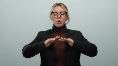 Sakin bir ifadeyle nefes alma egzersizi yapan kadın izole edilmiş beyaz arka planda gözlük takıyor ve siyah takım elbise giyiyor. Elleri birbirinin üstüne nazikçe yerleştirilmiş..