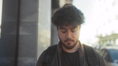 Güneşli bir günde açık havada gülümseyen, sıradan bir ceket ve sırt çantası giyen, yumuşak odaklı arka planı olan bir binanın yanında duran İspanyol asıllı genç bir adam..