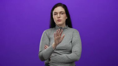 Canlı mor arka plana karşı hareket eden, gözlük takan ve gri başlıklı kadın, onaylanmamış ifadeler ve odaklanmış ifadelerle sözlü olmayan iletişim kuruyor..
