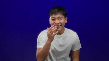 Mavi arka planda gülümseyen genç adam, eğlence ya da utanç ifade ediyor, günlük beyaz gömlek giyiyor..