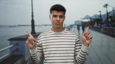 Genç adam deniz kenarındaki bir gezinti alanında sakin bir plaj geçmişine yansıyan rahat ve mizahi bir tavır sergiliyor..