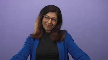 Middle aged hispanic woman wearing blue jacket and glasses extends open arms and gentle smile in purple studio; warmth.