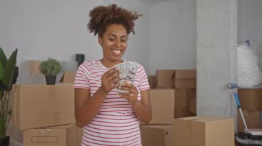Genç Afrikalı Amerikalı kadın yeni evdeki kutular arasında nakit para sayıyor; neşe dolu finansal başarı.