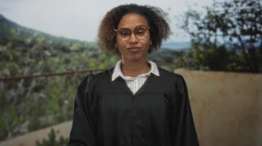 Cüppeli kadın yargıç gözlük takıyor, el kaldırıyor ve bina terasında saçlarına dokunuyor; alaycı şüphecilik.