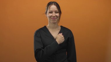 Yumruğunu sıkmış, öne doğru yumruk atan, gülümseyen, turuncu stüdyoda siyah bluz giyen, azimli genç İspanyol kadın..