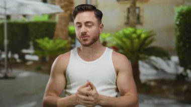 Sakallı ve beyaz atletli genç adam yeşilliklerle çevrili bir şehir caddesinde, düşünceli ve rahat görünüyor..