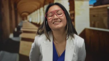 Gözlüklü bir kadın, kapalı bir üniversite koridorunda, mavi bir gömleğin üzerine beyaz ceket giyip, akademik bir ortamda pozitifliğini dile getiriyor..