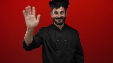 Sakallı İspanyol bir aşçı canlı kırmızı bir arkaplana el sallarken gülümsüyor. Karşılama ve profesyonel mutfak varlığını vurgulayan siyah bir üniforma giyiyor..