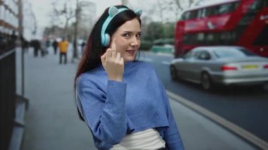 Kulaklıkla orta parmağını gösteren bir kadın. Arka planda otobüsle gülümseyen bir kadın. Mavi bir kazak giyiyor ve açık hava gününün tadını çıkarıyor..