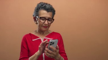 Stüdyo ortamında akıllı telefonu turuncu arka planda kullanan kadın, üst düzey kadın teknoloji etkileşimine odaklı modern iletişimi resmediyor.