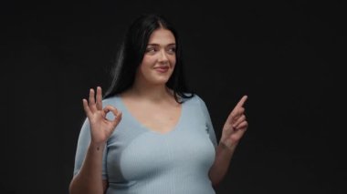 Artı boyunda bir kadın siyah bir arkaplana işaret ediyor, kendine güvenini ve pozitifliğini gülümseyerek ifade ediyor..