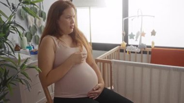 Eli ağzı kapalı hamile beyaz bir kadın stüdyo odasında çıplak karnını tutuyor..