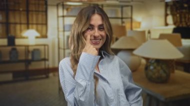 Bir mobilya galerisindeki lambalar arasında gülümserken işaret parmağını gözüne gösteren genç sarışın kadın, merak uyandıran bir oyunculuk..