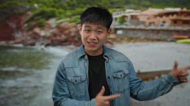 Kot ceketli genç adam arka planda canlı yeşil tepeler ve kırsal evler ile sahil manzarasını coşkuyla işaret ediyor..