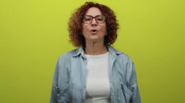 Orta yaşlı, esmer bir kadın canlı sarı arka plana karşı öpücük hareketi yapıyor. Günlük kıyafetler ve gözlük takıyor. Neşeli duygular ifade ediyor..