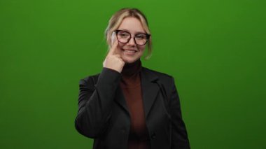 Siyah ceketli, yeşil arka planda gözlüklü, parlak renklerde bir stüdyo ortamında kendine güveni ve pozitifliği gösteren bir kadın..