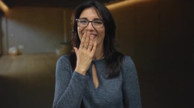 Orta yaşlı İspanyol kadın, hafif ışıklandırılmış stüdyo ortamında gülümserken ağzını eliyle kapatır..