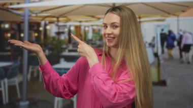 Dışarıdaki bir restoranda pembe bir gömlek giyerken terasta gülümseyen ve işaret eden bir kadın, canlı bir yemek sahnesi ve sıradan bir ortam sergiliyor..