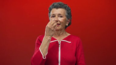 Kırmızı elbiseli, gri saçlı yaşlı bir kadın canlı kırmızı bir arkaplan karşısında el kol hareketi yapıyor, dört parmağı havada neşe ve güven ifadeleri sergiliyor..