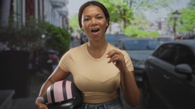 Pembe bir miğfer tutan ve sokakta sağ eliyle el kol hareketi yaparken gülümseyen bir kadın..