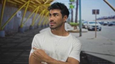 Beyaz tişörtlü genç adam park halindeki arabaların yanındaki güneşli şehir sokağında nesneyi işaret ediyor..