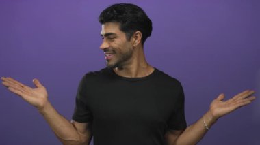 Genç İspanyol adam, mor stüdyo duvarına karşı avuçlarını açık bir şekilde hafifçe gülümseyerek omuz silker; belirsizlik.