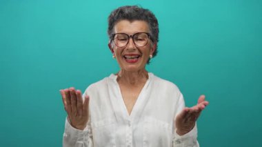Beyaz tişörtlü yaşlı bir kadın canlı mavi bir arkaplan karşısında sevinçle el kol hareketi yapıyor, gözlük takıyor ve sıcak bir şekilde gülümsüyor, dışavurumcu iletişim ve olumlu duygular sergiliyor..