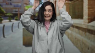 Esmer saçlı bir kadın, eski şehir sokaklarında kulak hareketi yapıyor. Gri kapüşonlu, neşeli bir şekilde gülümsüyor..