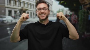 Sakallı ve gözlüklü genç adam bir şehir caddesinde başarıyı kutluyor, kendine güveni tam bir ifade sergiliyor..