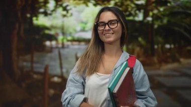 Gözlüklü, kırmızı sırt çantalı, elinde kitap olan ve kot pantolon ve beyaz tankla ağaçta sırıtan İspanyol bir kadın..