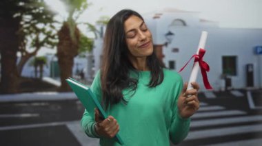 Elinde kırmızı kurdele ve deniz mavisi not defteri olan bir kadın, beyaz binalarla yaya caddesinde dikilirken gülümsüyor ve başını eğiyor; başarı gururu.