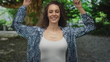 Kadın işaret parmağı parkın yukarısında yeşil ağaçların altında desenli gömlek ve beyaz akvaryum giyerek; sevinç iyimserliği güçlendirme.