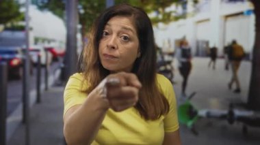 Sarı tişörtlü orta yaşlı İspanyol kadın, bulanık yoldan geçenlerin arasında hareketli şehir caddesini işaret ediyor; kararlılık özgüveni çözüyor.