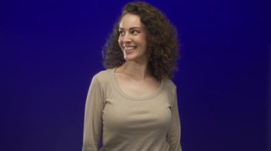 Stüdyoda koyu mavi arka plana bakarken bej rengi uzun kollu üst bluz giyen kadın gülümsüyor ve başparmağını kaldırıyor; özgüven teşviki.