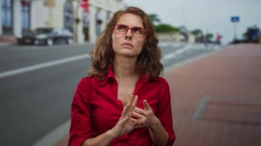 Kırmızı gömlekli, gözlüklü bir kadın şehir caddesinde düşünceli bir şekilde duruyor. Arka planda binalar, açık havada gündüz vakti bulanıklaşıyor..