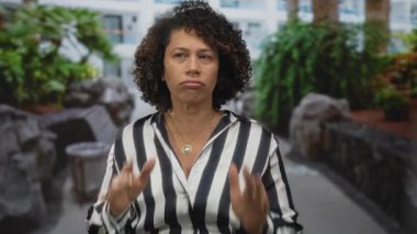 Orta yaşlı İspanyol kadın çizgili gömlek giyerek binanın dışında yumruklarını sıkar; kaygı dayanıklılığı kuvveti.