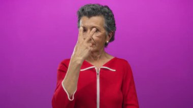 Kırmızı elbiseli yaşlı bir kadın canlı pembe bir arka plana karşı ihtiyatlı bir jest yapıyor..