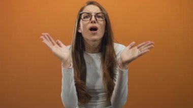 Woman with raised palms, open mouth and glasses in orange studio showing a startled shrug and wide eyes;surprise bewilderment frustration.