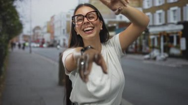 Beyaz t-shirt ve gözlük takmış, gülümseyen, neşeli bağlantı neşesi ile caddede kamerayı gösteren kadın parmağı.