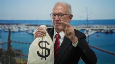 Takım elbiseli kıdemli bir adam elinde dolar işareti olan bir para çantası tutuyor. Arka planda botlarla deniz kenarında güvenle gülümsüyor ve yolsuzluğa işaret ediyor..